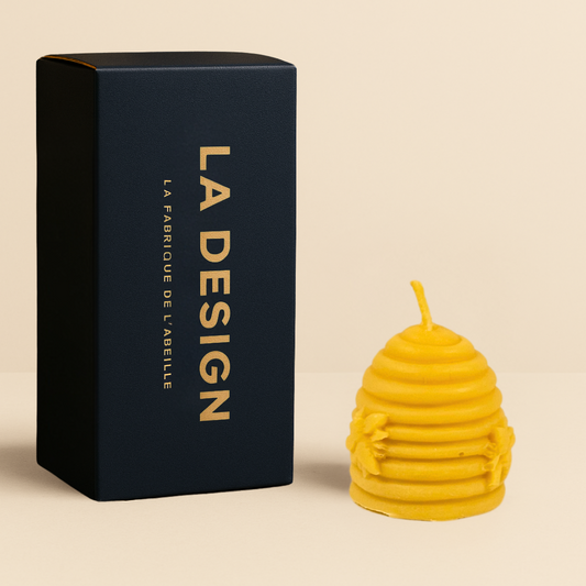 Bougie artisanale en cire d’abeille en forme de ruche, décorée d’abeilles en relief, accompagnée de son packaging bleu marine "LA DESIGN" doré, sur fond beige.