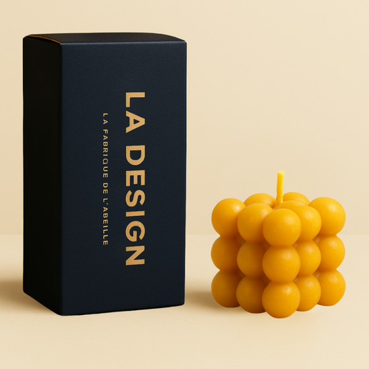 Bougie cube bulle jaune en cire d'abeille, accompagnée de son packaging bleu marine "LA DESIGN", sur fond beige chaleureux.