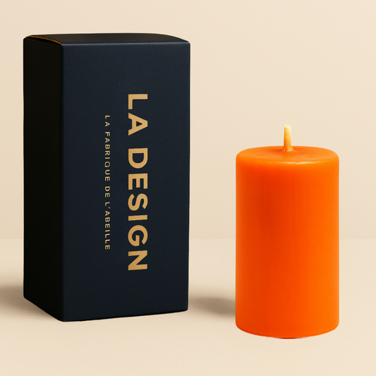 Bougie cylindrique orange avec packaging "LA DESIGN" bleu marine et doré de La Fabrique de l’Abeille, posée sur fond beige naturel.