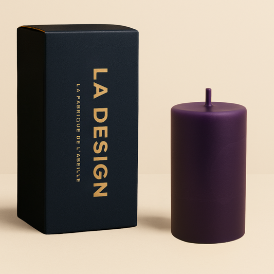 Bougie cylindrique violette accompagnée de son packaging bleu marine "LA DESIGN" doré, sur fond beige naturel.