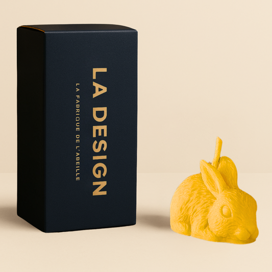 Bougie artisanale en forme de lapin, en cire d'abeille, accompagnée de son packaging bleu marine "LA DESIGN" doré, posée sur fond beige.