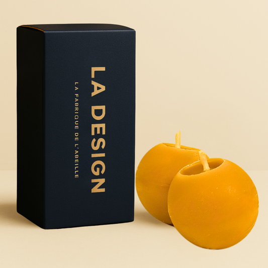 Duo de bougies artisanales en cire d'abeille, forme sphérique, accompagnées de leur packaging bleu marine "LA DESIGN" doré, posées sur un fond beige.